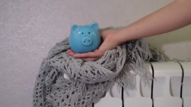 Mavi domuzcuk bankası elektrik ve ısıtma masraflarından tasarruf ediyor, kapat. Tasarruf kavramı. Kadın, kumbaraya bozuk para koyuyor ve radyatörün üzerinde duruyor..