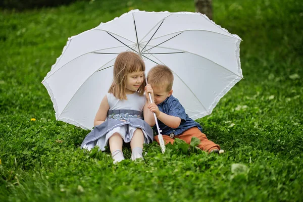 어린 소녀와 소년은 우산 밑에 숨어 있습니다. 형 과 누나 가 공원에서 놀고 있어. — 스톡 사진