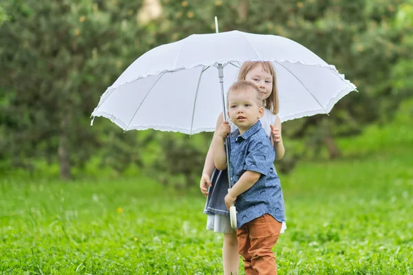 Rapariga e rapaz estão escondidos debaixo de um guarda-chuva. irmão e irmã estão brincando no parque. — Fotografia de Stock
