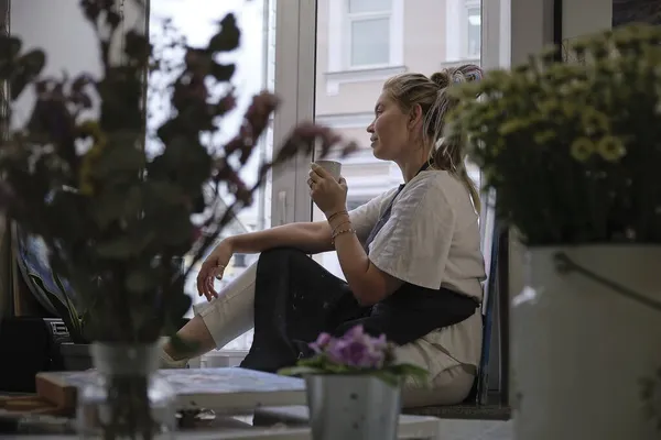 Kirli önlüklü sanatçı kadın bir fincan çayla pencerenin kenarında oturuyor ve pencereden dışarı bakıyor. — Stok fotoğraf