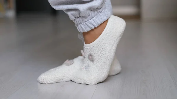 Gambe di una donna in calzini bianchi che cammina sul pavimento di legno della sua casa con un divano sullo sfondo. piedi con calzini bianchi sul pavimento grigio in legno — Foto Stock