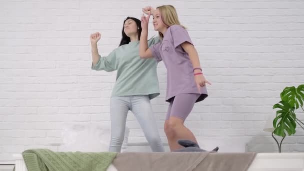 Chicas jóvenes bailando en la cama en casa. Dos jóvenes multinacionales pasando tiempo juntas, pijama party. mejores novias tomar selfies. — Vídeos de Stock
