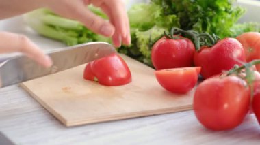 Mutfak bıçağını kullanarak ahşap kesim tahtasıyla taze domates, domates filizi ve arka planda marul kesiyor.