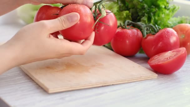 Hands using kitchen knife cutting fresh tomato on wooden cutting board, with sprig of tomatoes and lettuce on background — Video Stock