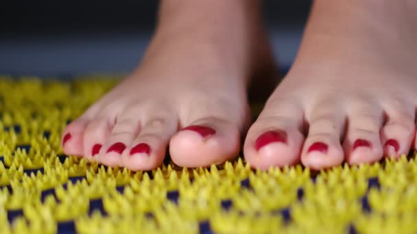 圧力マットの上に立つ白人女性の足を持つ自家製鍼治療マッサージ。黄色のプラスチック製のピンを持つ青いマットレスの上の女性の足の正面図 — ストック動画