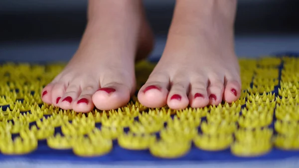 Домашний акупунктурный массаж с кавказскими женскими ногами, стоящими на коврике с акупрессурой. передний вид женских ног на синем матрасе с желтыми пластиковыми булавками — стоковое фото