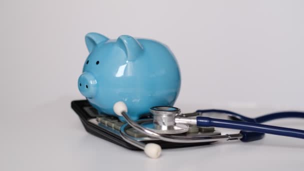 Hucha con estetoscopio y calculadora sobre fondo blanco, concepto de seguro de salud. El costo de la medicina. — Vídeos de Stock