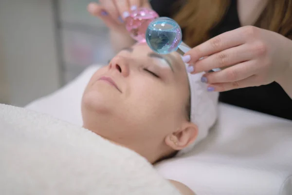 Paciente calma mulher submetida aos procedimentos de massagem facial cosmética para rejuvenescimento da pele rosto e médico usando instrumentos cosméticos, bolas de gelo massagem. — Fotografia de Stock