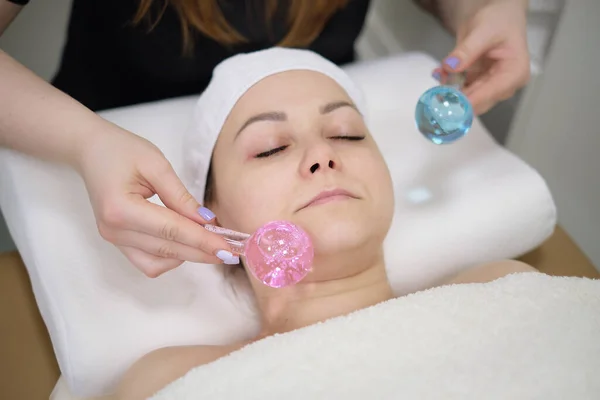 Paciente calma mulher submetida aos procedimentos de massagem facial cosmética para rejuvenescimento da pele rosto e médico usando instrumentos cosméticos, bolas de gelo massagem. — Fotografia de Stock