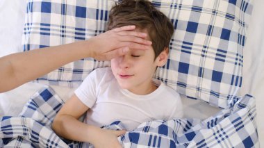 Hasta oğlunun alnında başını tutan şefkatli bir anne. Çocuk sağlığı konsepti. Üzgün küçük çocuk yatakta yatıyor..