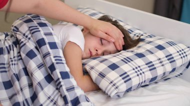 Hasta oğlunun alnında başını tutan şefkatli bir anne. Çocuk sağlığı konsepti. Üzgün küçük çocuk yatakta yatıyor..