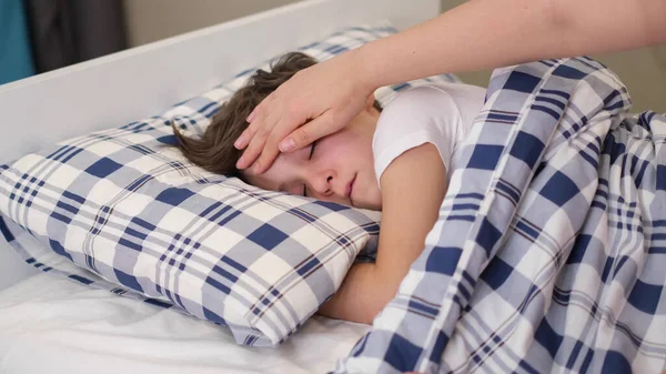 caring mother holding head on sick son forehead. Children healthcare concept. Sad little boy lies on the bed.