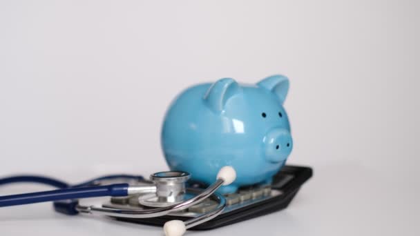 Банк свиней со стетоскопом и калькулятором на белом фоне, концепция медицинского страхования — стоковое видео