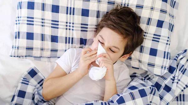 Enfermedad del resfriado infantil tejido que sopla secreción nasal. niño pequeño está acostado en la cama y sonando la nariz en papel de seda en casa. Alergia, gripe —  Fotos de Stock