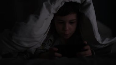 Karanlıkta telefonla oynayan çocuk. Uzun süreli telefon oyunları küçük çocukların görme ve sağlığını olumsuz etkiler. Cep telefonuna bağımlılık.