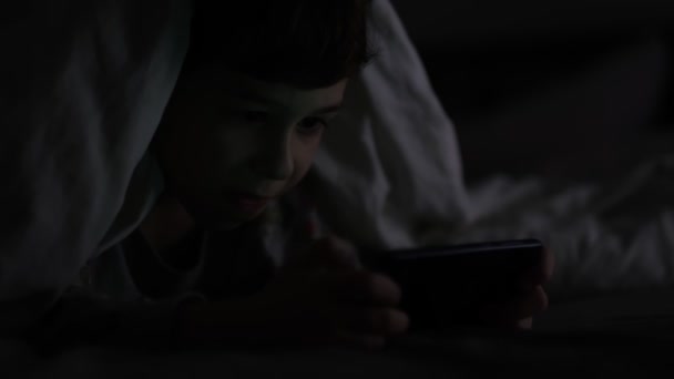 Een jongen die in het donker aan de telefoon speelt. Langdurig telefoneren heeft negatieve gevolgen voor het gezichtsvermogen en de gezondheid van jonge kinderen. Afhankelijkheid van een mobiele telefoon. — Stockvideo