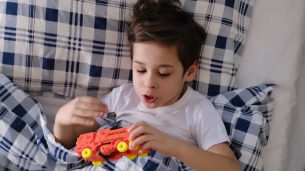 A kisfiú az ágyban fekszik, takaróval borítva, és a narancssárga műanyag robotjával játszik. a gyermek felépült a betegségből, és jó hangulatban van, — Stock videók