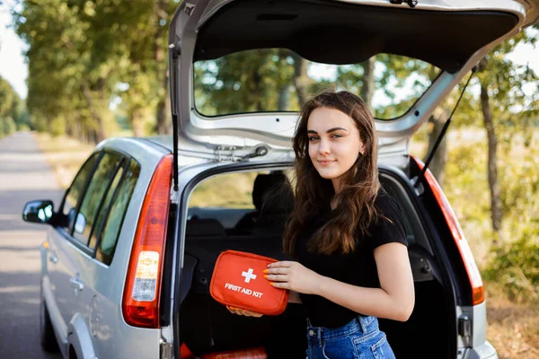 Gümüş hatchback arabasının arka kapısının yanında duran ve acil durumlar için her arabada olması gereken ilk yardım çantasını gösteren genç bir kız.