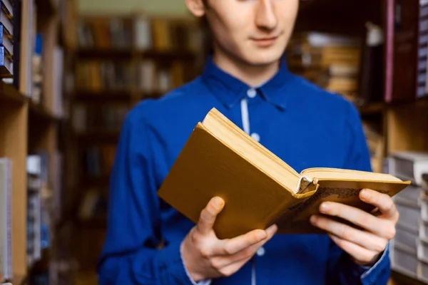Close up picture of a book in male student`s hands standing between shelves of books in the library