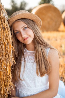 Hasır şapkalı, beyaz elbiseli çekici bir kızın saman balyasına yaslanmış portresi.