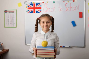 Bir yığın kitabı ve sınıfın beyaz tahtasının yanında bir elması olan mutlu okul kızı. Öğrenci kız karantina ve uzaktan öğrenme sürecinden sonra okula döndüğü için mutlu.