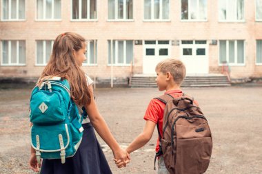 Okul çantalı iki mutlu öğrencinin el ele tutuşup okula yürüdüğü arka plan. Arkadaşlar yaz tatilinden sonraki ilk günlerinden bahsederler..