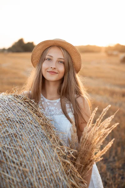 Beyaz elbiseli ve hasır şapkalı çekici bir genç kız elinde bir buket kurutulmuş tarla çiçeği tutan saman balyasının yanında duruyor. Hasat kavramı, sonbahar
