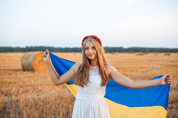 Smiling Ukrainian girl in white dress and red hair hoop holding blue and yellow flag. Patriotic girl in agricultural field after the harvest. Ukraine's independence day. National flag day of Ukraine.