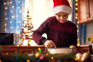 Noel şapkalı genç bir çocuk arka planda süslenmiş köknar ağacıyla süslenmiş ışıklarla dolu bir mutfakta annesine kurabiye pişirmesinde yardım eder. Tatil hazırlığı, aile zamanı..