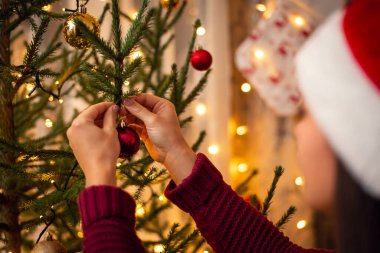 Köknar ağacının yanında dalına kırmızı bir mücevher asan genç bir kadın resmi. Noel zamanı, neşeli atmosfer, mucizeleri bekliyor..