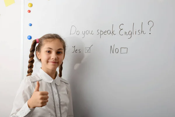 Little smiling girl in school uniform near the board in the classroom. Showing thumbs up, happy to know English, speak foreign language