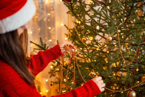크리스마스 모자를 스웨터를 갈색의 소녀가 구슬로 전나무 장식을 뒤에서 보인다 — 스톡 사진