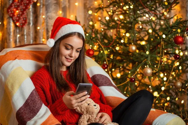 Noel, çevrimiçi yaşam kavramları, sıcak renkler, şenlikli atmosfer. Neşeli, güzel bir kız koltukta oturuyor, elinde cep telefonu, arkadaşlarıyla sohbet ediyor..