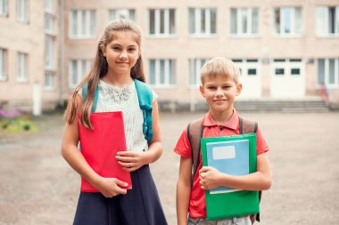 Okul yakınlarında öğrenim materyalleri ve sırt çantalarıyla ders çalışmaya hazır, kameraya gülümsüyor.