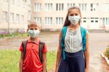Tıp maskeli mutsuz okul çocukları. Oğlan ve kız sırt çantalarını taşıyarak okulun yanında durup kameraya bakıyorlar. Coronavirus karantinası ve tecritten sonra okula dönüyorum..