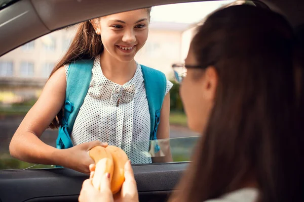 Smiling girl takes lunch from caring mother, who is sitting in car and giving instructions to her daughter before the first day at school after summer holiday. Back to school, healthy nutrition.