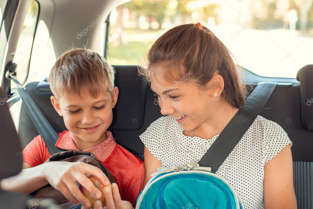 Two siblings sit on the back sits of the car, fastened with seat belts, hold their backpacks and cheerfully play with an apple on their way to school.