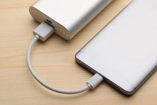 Silver smartphone charging with a power bank on a light wooden background