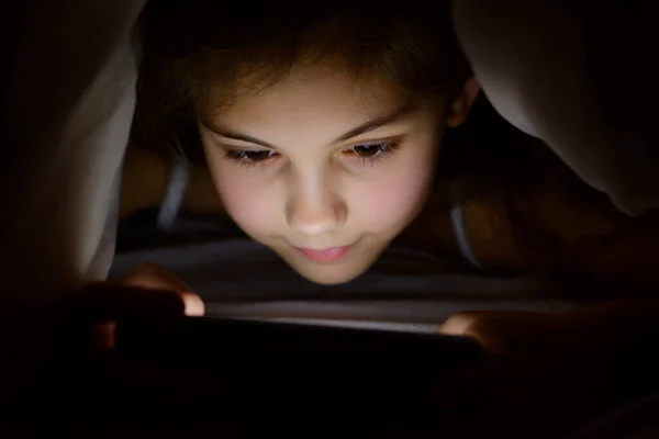 Beautiful caucasian girl playing games on her mobile phone under the blanket at night. Little child don`t want to sleep and plays on smartphone under the cover in order parents don`t see her