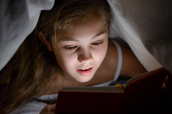 Impressed little girl under the blanket with a book and a flashlight reading horrors before go to bed. Small female child covered with blanket and enjoy reading scary stories in her bed at night