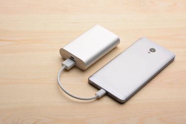 Modern gümüş akıllı telefon elektrik bankasıyla şarj edilirken arka paneli açık, açık ahşap masanın üzerinde yatıyor.