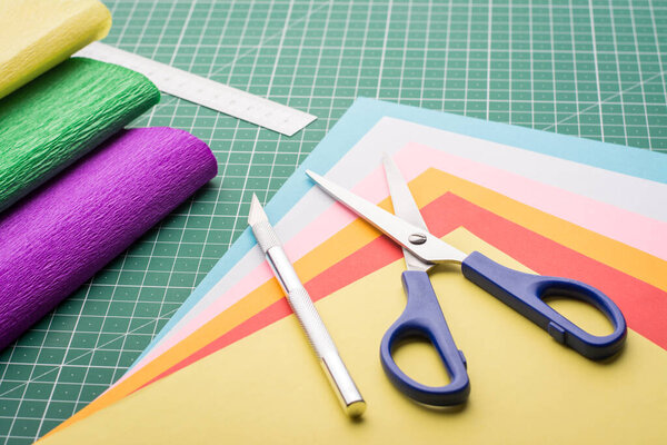 Комплект различного стационарного оборудования и разноцветной бумаги на коврике для резки. Ножницы, бумажный нож и линейка на коврике для резки