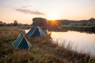 Hafta sonlarını nehir kıyısında geçirmek, çadırda yaşamak. Nehrin kenarında iki çadır var. Arka planda yükselen güneş var.