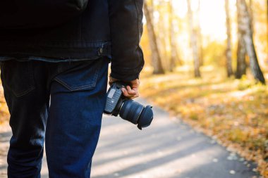 Fotoğrafçının elindeki kameranın yakın çekim görüntüsü. Sırtından fotoğrafçı, elinde kamerayla parkta yürüyor. Arka planda sonbahar görünümü.