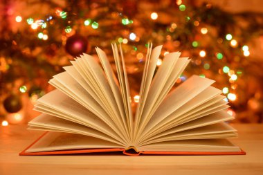 Christmas time, open book of fairy tales on the table, christmas tree with colorful lights and decoration on the background. Warm colours and festive atmosphere.