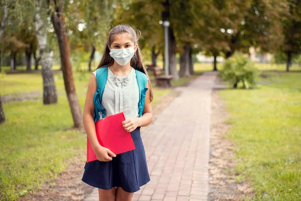 Schoolgirl in protective medical mask, wearing backpack and holding red folder with materials going to school through the park