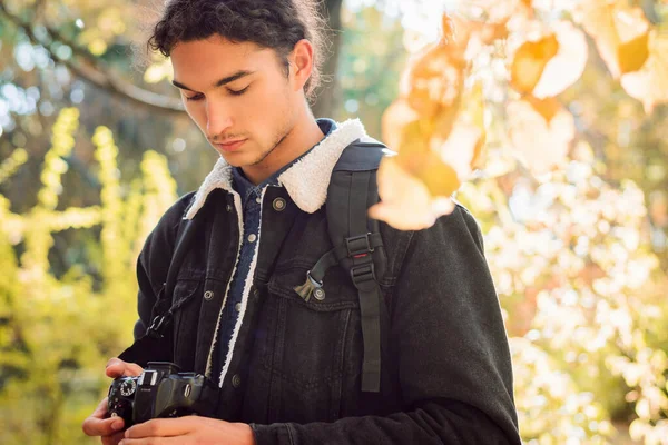 Handsome young man standing in the forest on a bright sunny autumn day, looking at his camera after taking beautiful pictures of the trees and golden foliage.