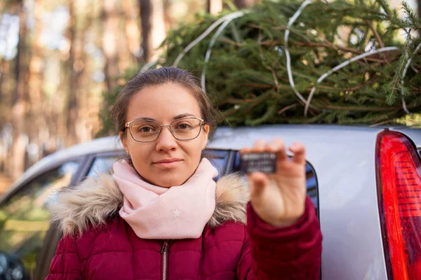 Young girl in glasses standing near the car with a christmas tree on its roof and showing a chip, which she has got after buying a tree. Legal shopping for winter holidays.