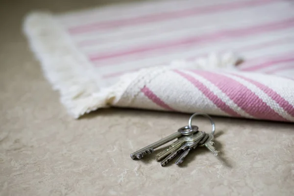 Close-up photo of keys, hidden under the mat near the entrance of the house or flat. Grey floor and thin striped carpet.