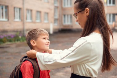 Mother saying goodbye to her little son, adjusting collar of his shirt and telling him to behave nice in school